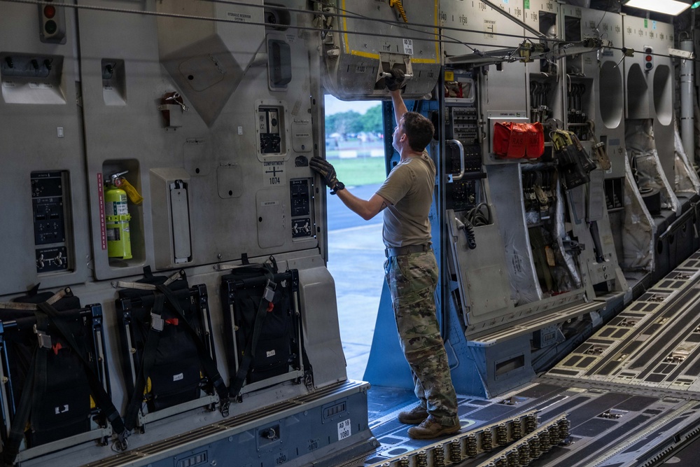 A military service member in camouflage uniform is working on the interior wall panel of a large military cargo aircraft, likely a C-17 Globemaster III. The person is standing on the cargo floor near an open side door, reaching upward to operate or inspect overhead mechanisms. The interior shows complex aircraft systems panels, safety equipment, and cargo management hardware.