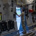 A military service member in camouflage uniform is working on the interior wall panel of a large military cargo aircraft, likely a C-17 Globemaster III. The person is standing on the cargo floor near an open side door, reaching upward to operate or inspect overhead mechanisms. The interior shows complex aircraft systems panels, safety equipment, and cargo management hardware.