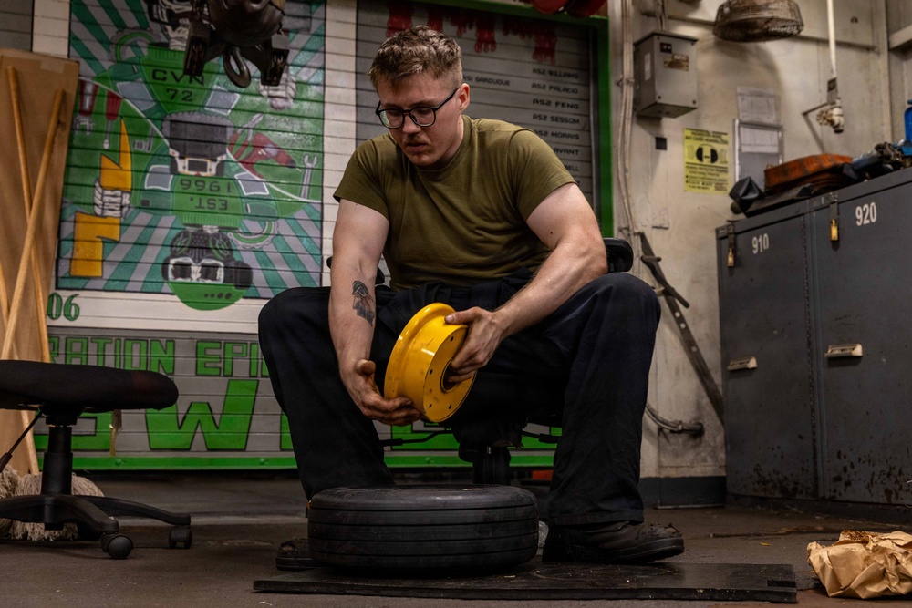 A young male in a military work environment is seated and manually assembling or disassembling a yellow wheel hub onto a black tire. The setting appears to be a maintenance shop aboard a U.S. Navy aircraft carrier, indicated by the 'CVN 72' marking visible on the mural in the background. The worker is wearing an olive green t-shirt, black pants, and glasses, with a visible tattoo on his forearm.