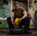 A young male in a military work environment is seated and manually assembling or disassembling a yellow wheel hub onto a black tire. The setting appears to be a maintenance shop aboard a U.S. Navy aircraft carrier, indicated by the 'CVN 72' marking visible on the mural in the background. The worker is wearing an olive green t-shirt, black pants, and glasses, with a visible tattoo on his forearm.