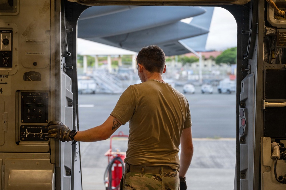 A military personnel member in olive/tan uniform and multicam pants stands at an open aircraft door, looking out onto a flight line or tarmac. The individual is positioned inside what appears to be a large military transport aircraft, holding onto the door frame with a gloved hand. The setting appears to be a tropical or subtropical airfield based on the visible vegetation and architecture in the background.