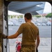 A military personnel member in olive/tan uniform and multicam pants stands at an open aircraft door, looking out onto a flight line or tarmac. The individual is positioned inside what appears to be a large military transport aircraft, holding onto the door frame with a gloved hand. The setting appears to be a tropical or subtropical airfield based on the visible vegetation and architecture in the background.