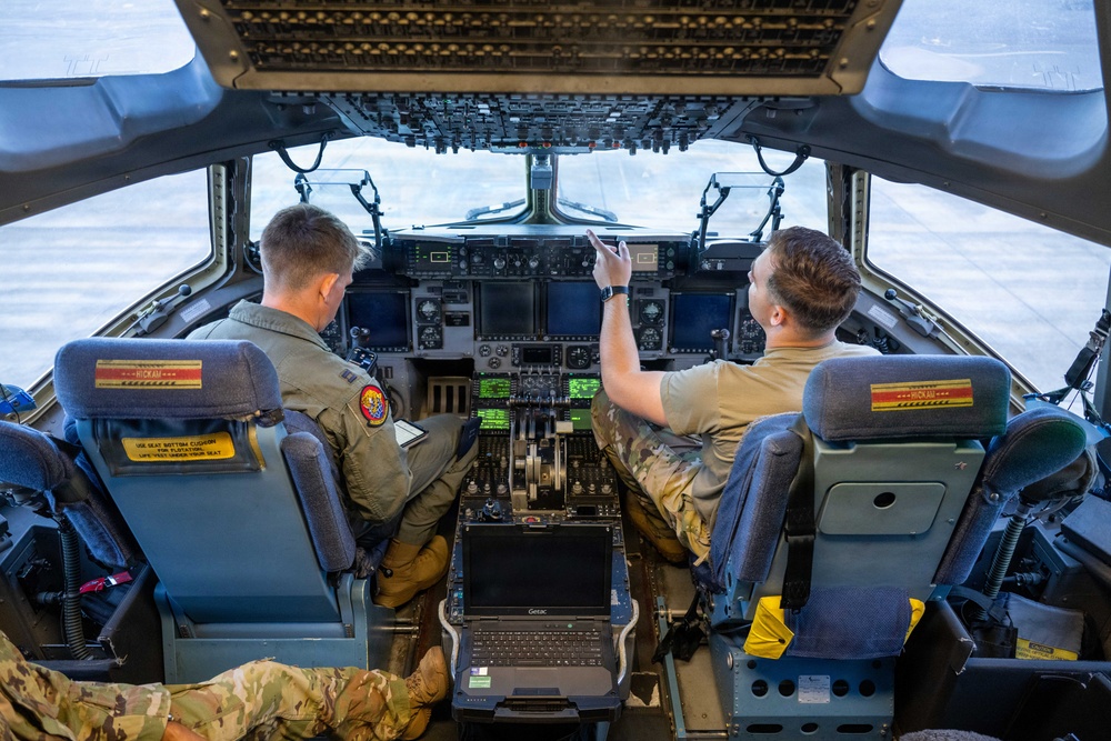 Two military aircrew members are seated in the cockpit of a large military transport aircraft, likely a C-17 Globemaster III, conducting pre-flight or ground operations checks. One crew member in a flight suit is reviewing documents while the other in a tan uniform is pointing toward the instrument panel or windscreen. The aircraft appears to be stationary on a flight line or tarmac.