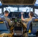 Two military aircrew members are seated in the cockpit of a large military transport aircraft, likely a C-17 Globemaster III, conducting pre-flight or ground operations checks. One crew member in a flight suit is reviewing documents while the other in a tan uniform is pointing toward the instrument panel or windscreen. The aircraft appears to be stationary on a flight line or tarmac.