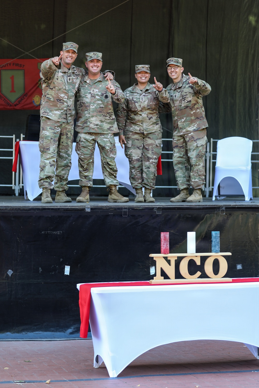 Four U.S. Army soldiers in OCP (Operational Camouflage Pattern) uniforms stand on a stage, posing with index fingers raised in a celebratory gesture. The setting appears to be an NCO (Non-Commissioned Officer) ceremony or induction event, evidenced by the 'NCO' wooden sign on the table in the foreground and the 1st Infantry Division banner visible in the background. The soldiers are smiling and appear to be celebrating a milestone occasion.