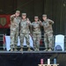 Four U.S. Army soldiers in OCP (Operational Camouflage Pattern) uniforms stand on a stage, posing with index fingers raised in a celebratory gesture. The setting appears to be an NCO (Non-Commissioned Officer) ceremony or induction event, evidenced by the 'NCO' wooden sign on the table in the foreground and the 1st Infantry Division banner visible in the background. The soldiers are smiling and appear to be celebrating a milestone occasion.