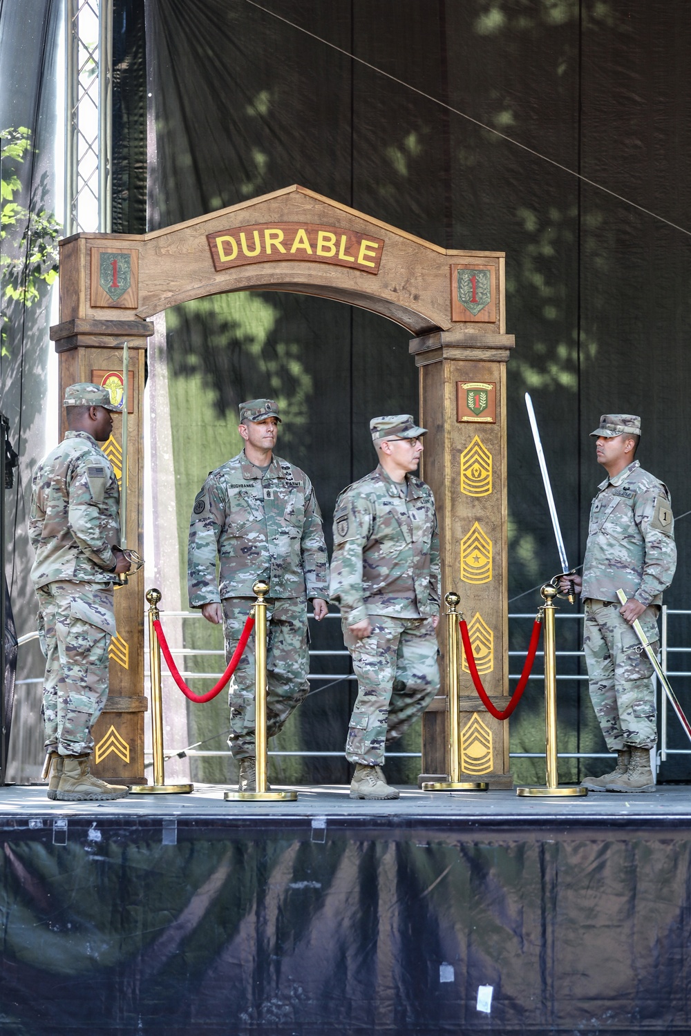 A formal U.S. Army military ceremony is taking place on a stage featuring a wooden arch inscribed with the word 'DURABLE' and decorated with unit insignia patches. Four soldiers in OCP (Operational Camouflage Pattern) uniforms are positioned on stage, two of whom are holding ceremonial sabers. Red velvet rope stanchions with gold posts frame the ceremonial arch area.