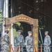 A formal U.S. Army military ceremony is taking place on a stage featuring a wooden arch inscribed with the word 'DURABLE' and decorated with unit insignia patches. Four soldiers in OCP (Operational Camouflage Pattern) uniforms are positioned on stage, two of whom are holding ceremonial sabers. Red velvet rope stanchions with gold posts frame the ceremonial arch area.