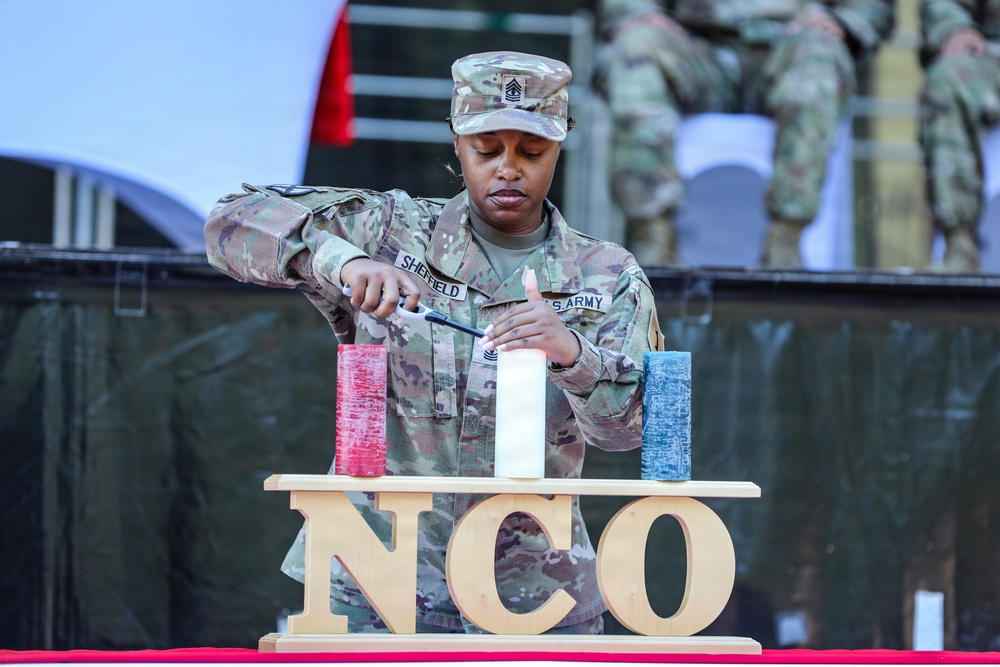 A U.S. Army NCO (Non-Commissioned Officer) identified by name tape as 'Sheffield' is lighting candles during what appears to be an NCO induction or recognition ceremony. Three candles in red, white, and blue are displayed on a wooden stand bearing the letters 'NCO'. The soldier wears the rank of First Sergeant based on the chevrons visible on her patrol cap.