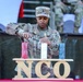 A U.S. Army NCO (Non-Commissioned Officer) identified by name tape as 'Sheffield' is lighting candles during what appears to be an NCO induction or recognition ceremony. Three candles in red, white, and blue are displayed on a wooden stand bearing the letters 'NCO'. The soldier wears the rank of First Sergeant based on the chevrons visible on her patrol cap.