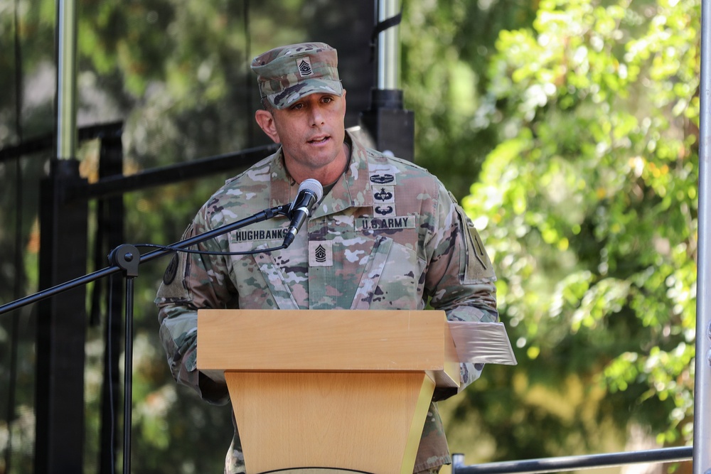 A U.S. Army soldier identified by name tape as 'HUGHBANKS' is speaking at a podium with a microphone. He is wearing the Operational Camouflage Pattern (OCP) uniform with visible rank insignia indicating a senior non-commissioned officer, along with qualification badges. The setting appears to be an outdoor military ceremony or official event.