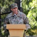A U.S. Army soldier identified by name tape as 'HUGHBANKS' is speaking at a podium with a microphone. He is wearing the Operational Camouflage Pattern (OCP) uniform with visible rank insignia indicating a senior non-commissioned officer, along with qualification badges. The setting appears to be an outdoor military ceremony or official event.