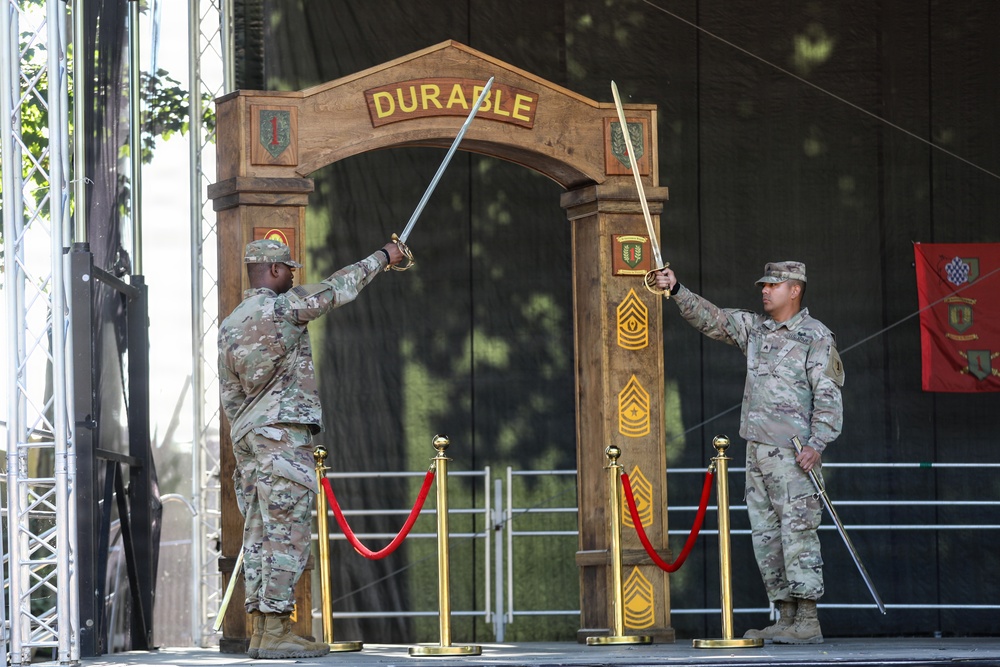 Two U.S. Army soldiers in OCP (Operational Camouflage Pattern) uniforms are crossing swords in front of a ceremonial wooden arch labeled 'DURABLE' on a stage. The arch displays various military rank insignia and unit patches, suggesting this is a formal military ceremony, likely a promotion, change of command, or unit activation event. Gold stanchions with red rope barriers frame the ceremonial arch.