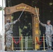 Two U.S. Army soldiers in OCP (Operational Camouflage Pattern) uniforms are crossing swords in front of a ceremonial wooden arch labeled 'DURABLE' on a stage. The arch displays various military rank insignia and unit patches, suggesting this is a formal military ceremony, likely a promotion, change of command, or unit activation event. Gold stanchions with red rope barriers frame the ceremonial arch.