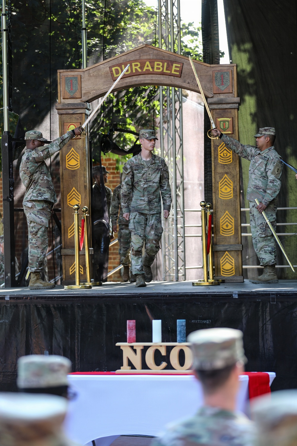 A U.S. Army NCO induction ceremony is taking place on a decorated stage. A soldier walks through an ornamental arch labeled 'DURABLE' while two soldiers on either side hold crossed sabers overhead in a traditional sword arch formation. The foreground shows an 'NCO' wooden sign on a table, with an audience of uniformed soldiers watching.