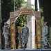 A U.S. Army NCO induction ceremony is taking place on a decorated stage. A soldier walks through an ornamental arch labeled 'DURABLE' while two soldiers on either side hold crossed sabers overhead in a traditional sword arch formation. The foreground shows an 'NCO' wooden sign on a table, with an audience of uniformed soldiers watching.