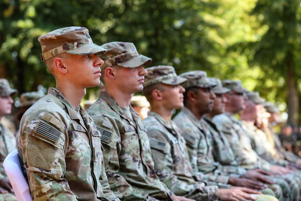 A row of U.S. Army soldiers in Operational Camouflage Pattern (OCP) uniforms are seated in formation, likely attending a military ceremony or formal event outdoors. The soldiers are seated in profile view, facing the same direction with attentive postures. The setting appears to be a tree-lined outdoor venue during daylight hours.