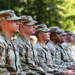 A row of U.S. Army soldiers in Operational Camouflage Pattern (OCP) uniforms are seated in formation, likely attending a military ceremony or formal event outdoors. The soldiers are seated in profile view, facing the same direction with attentive postures. The setting appears to be a tree-lined outdoor venue during daylight hours.