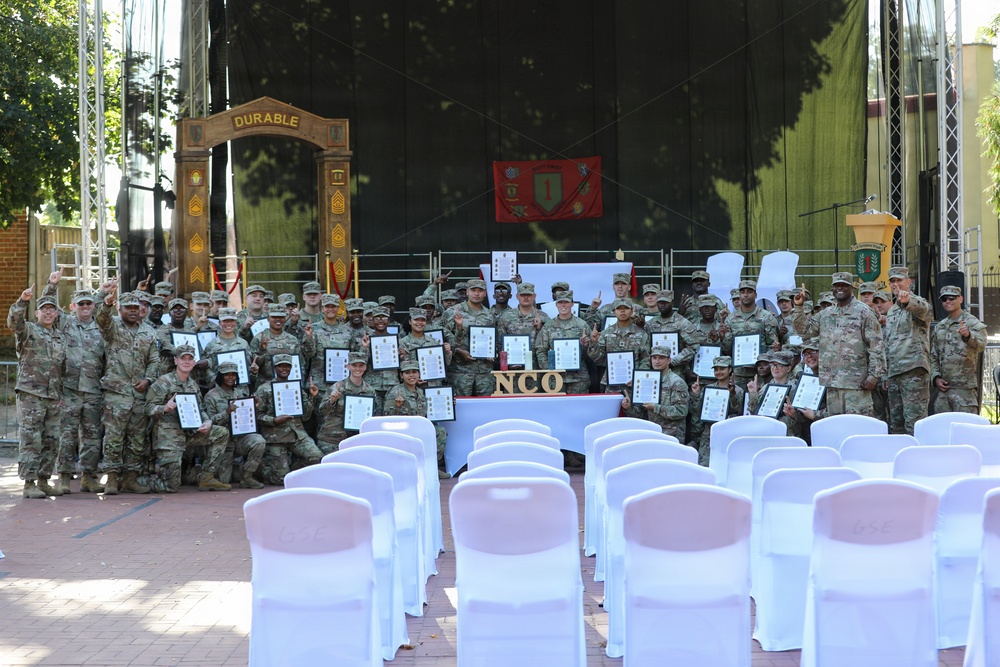 A large group of U.S. Army soldiers in camouflage uniforms pose for a group photo at what appears to be an NCO (Non-Commissioned Officer) induction or graduation ceremony. Many soldiers are holding framed certificates and raising index fingers in a celebratory gesture. The event takes place on an outdoor stage area decorated with a 'DURABLE' arch, military guidons, and white chairs arranged in rows.