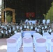 A large group of U.S. Army soldiers in camouflage uniforms pose for a group photo at what appears to be an NCO (Non-Commissioned Officer) induction or graduation ceremony. Many soldiers are holding framed certificates and raising index fingers in a celebratory gesture. The event takes place on an outdoor stage area decorated with a 'DURABLE' arch, military guidons, and white chairs arranged in rows.