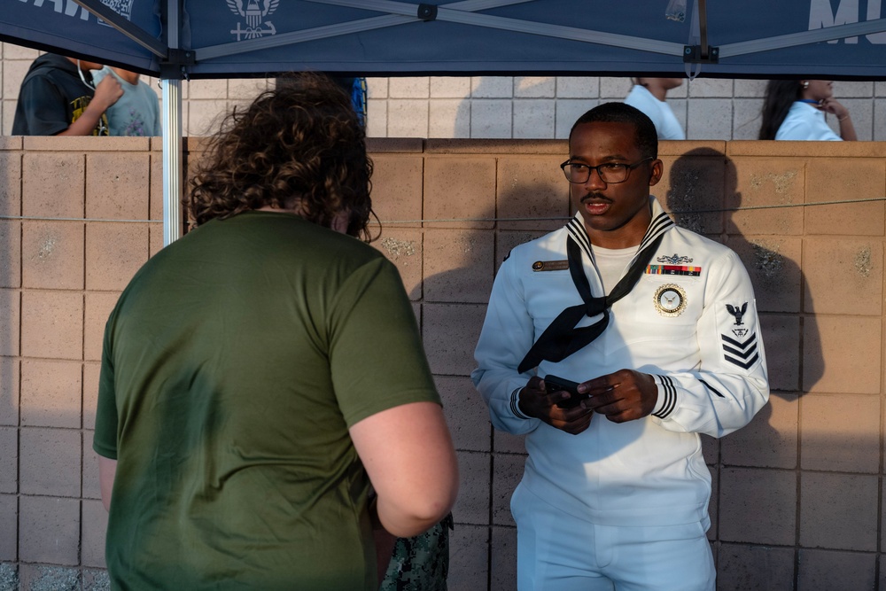 A U.S. Navy sailor in dress white uniform is engaged in conversation with a civilian wearing a green t-shirt at what appears to be an outdoor recruiting or community event. The sailor is wearing a Petty Officer First Class rank insignia, service ribbons, and a black neckerchief, and appears to be holding a mobile device. The setting includes a blue canopy tent with a military emblem visible overhead.