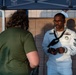 A U.S. Navy sailor in dress white uniform is engaged in conversation with a civilian wearing a green t-shirt at what appears to be an outdoor recruiting or community event. The sailor is wearing a Petty Officer First Class rank insignia, service ribbons, and a black neckerchief, and appears to be holding a mobile device. The setting includes a blue canopy tent with a military emblem visible overhead.