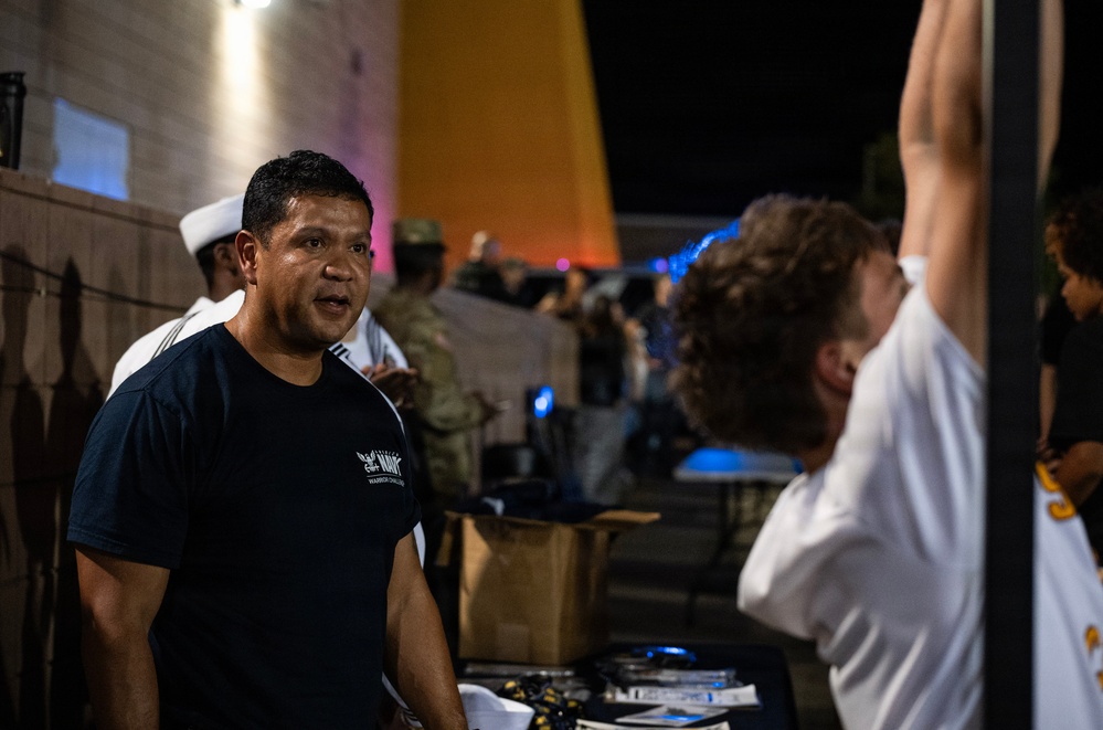 A nighttime outdoor event featuring military personnel and civilians gathered around what appears to be a recruitment or information table. A man in a dark Navy 'Warrior Challenge' t-shirt is speaking, while another person in a white shirt has their arm raised, possibly being measured or assessed. Military personnel in uniform, including a sailor in a white cover (hat), are visible in the background.