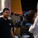 A nighttime outdoor event featuring military personnel and civilians gathered around what appears to be a recruitment or information table. A man in a dark Navy 'Warrior Challenge' t-shirt is speaking, while another person in a white shirt has their arm raised, possibly being measured or assessed. Military personnel in uniform, including a sailor in a white cover (hat), are visible in the background.