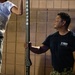 A man wearing a Navy Warrior Challenge t-shirt and camouflage pants holds a 'BULLBAR' pull-up bar while observing a younger person performing or preparing to perform an exercise on it. The setting appears to be an outdoor or semi-outdoor area with a concrete block wall in the background. The scene suggests a military recruitment or fitness demonstration event.