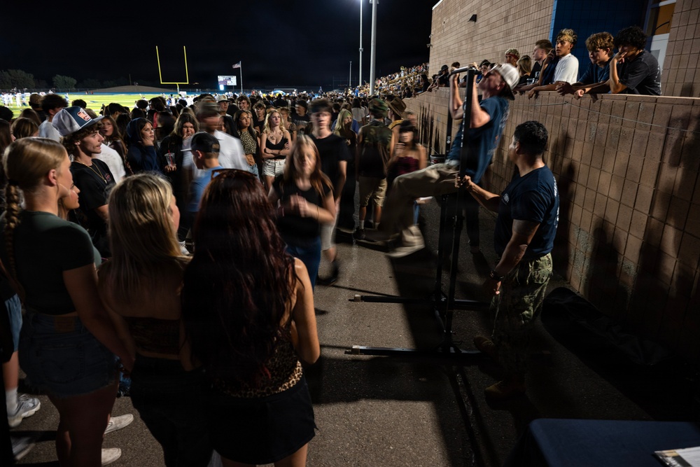 A large crowd of young people, likely high school or college students, gathered at an outdoor football stadium at night. The scene appears to show spectators near a concrete block wall separating a lower walkway from elevated bleachers, with some individuals climbing or sitting on the wall. A football field with goal posts and stadium lights is visible in the background.
