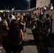 A large crowd of young people, likely high school or college students, gathered at an outdoor football stadium at night. The scene appears to show spectators near a concrete block wall separating a lower walkway from elevated bleachers, with some individuals climbing or sitting on the wall. A football field with goal posts and stadium lights is visible in the background.