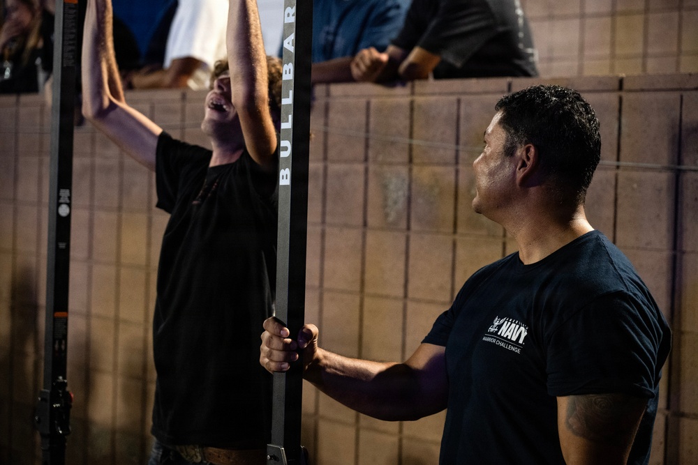 Two men are engaged in a physical fitness challenge, with one man performing a pull-up or hang on a 'Bullbar' piece of equipment while the other, wearing a 'Navy Warrior Challenge' t-shirt, appears to be spotting or officiating. The setting appears to be an outdoor or semi-outdoor event with spectators visible in the background leaning over a wall. The scene suggests a military recruitment or fitness competition event.