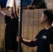 Two men are engaged in a physical fitness challenge, with one man performing a pull-up or hang on a 'Bullbar' piece of equipment while the other, wearing a 'Navy Warrior Challenge' t-shirt, appears to be spotting or officiating. The setting appears to be an outdoor or semi-outdoor event with spectators visible in the background leaning over a wall. The scene suggests a military recruitment or fitness competition event.