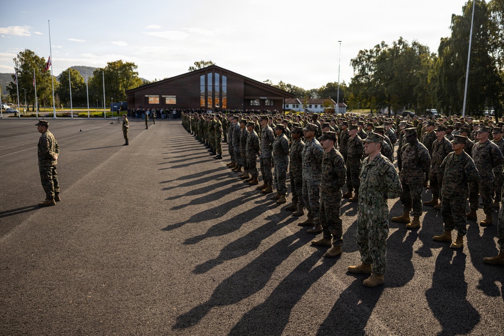 A large formation of military personnel, predominantly in U.S. Marine Corps MARPAT woodland camouflage uniforms, are standing at attention on a parade ground or drill deck. At least one individual in Navy Working Uniform (NWU Type III) is visible in the foreground, suggesting a joint-service formation. The setting appears to be a military installation with a brick building, flagpoles, and tree-lined hills in the background.