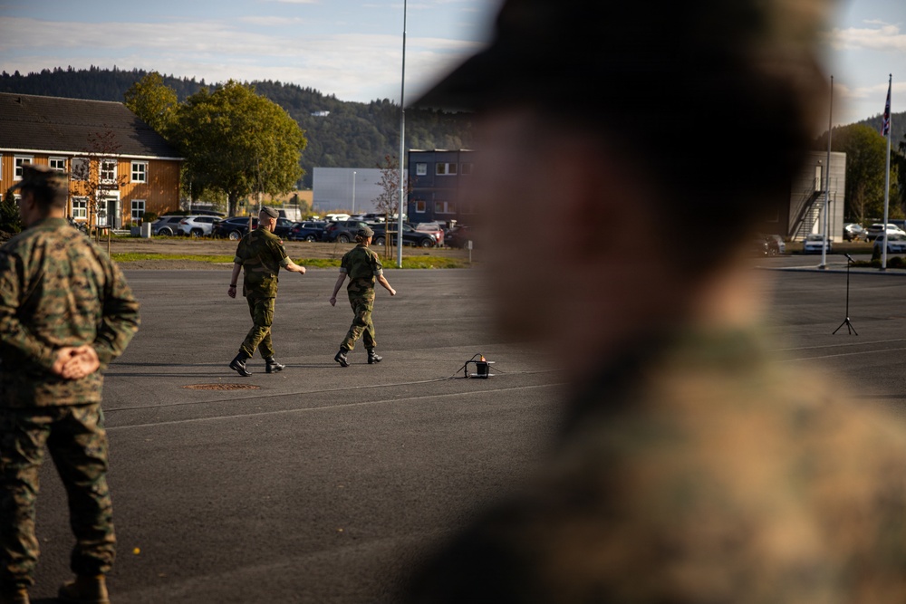 The image shows a military parade ground or assembly area with personnel in different camouflage uniforms. Two soldiers in what appears to be Norwegian/European-style camouflage are walking across a paved area, while figures in what appears to be US Marine Corps MARPAT camouflage are visible in the foreground. The setting appears to be a military installation with buildings, parked vehicles, trees, and a flagpole visible in the background.