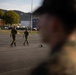 The image shows a military parade ground or assembly area with personnel in different camouflage uniforms. Two soldiers in what appears to be Norwegian/European-style camouflage are walking across a paved area, while figures in what appears to be US Marine Corps MARPAT camouflage are visible in the foreground. The setting appears to be a military installation with buildings, parked vehicles, trees, and a flagpole visible in the background.