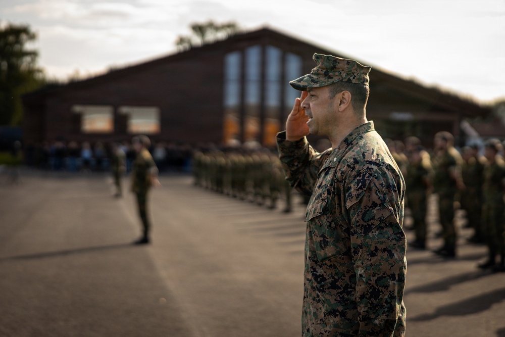 A military officer in digital camouflage uniform renders a salute during what appears to be a formal parade or ceremony. Rows of uniformed personnel are standing at attention in formation on a paved area, with a large brick building visible in the background. The image is captured during golden hour lighting, giving it a warm, dramatic tone.