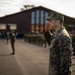 A military officer in digital camouflage uniform renders a salute during what appears to be a formal parade or ceremony. Rows of uniformed personnel are standing at attention in formation on a paved area, with a large brick building visible in the background. The image is captured during golden hour lighting, giving it a warm, dramatic tone.