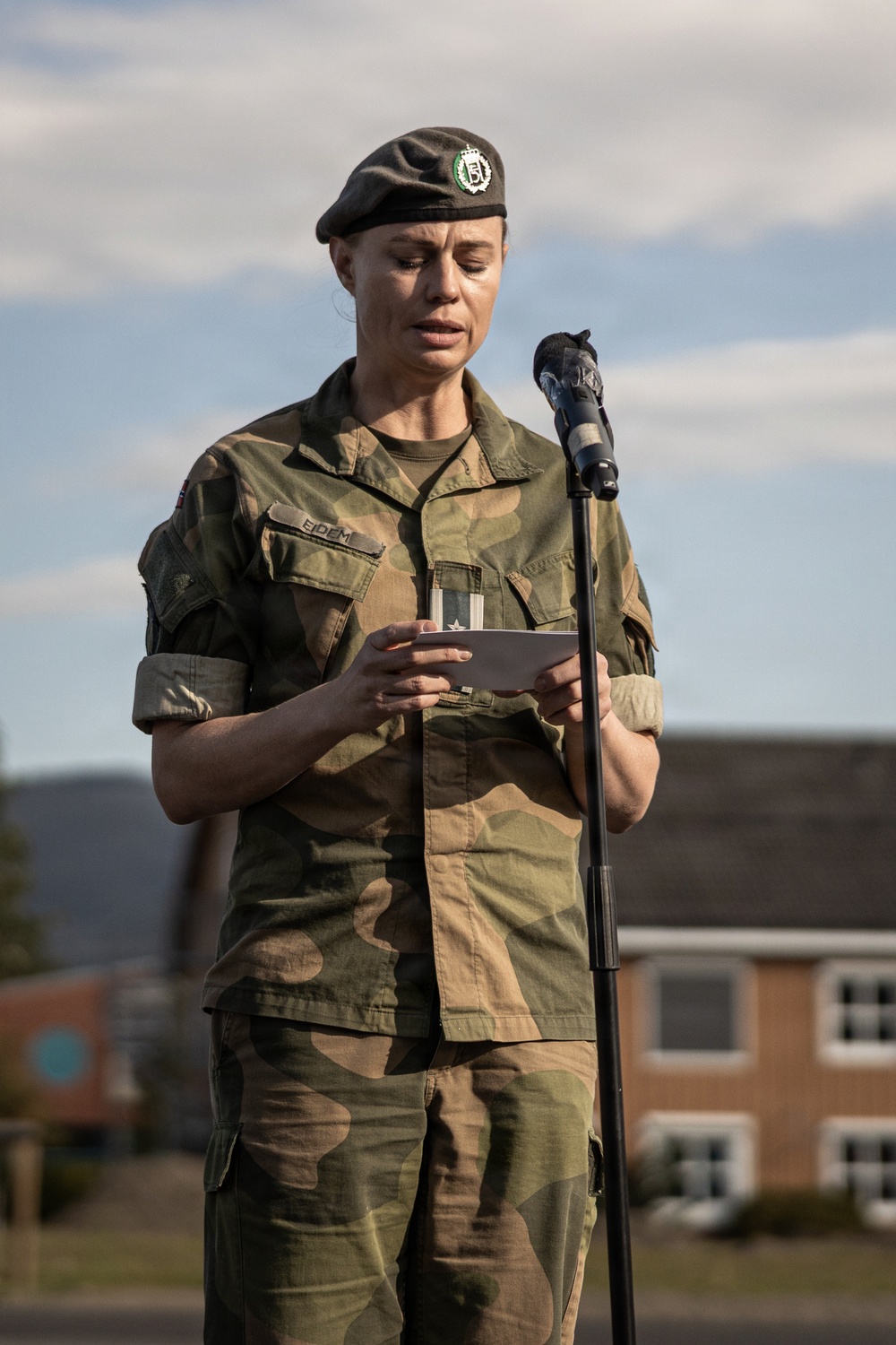 A female military officer in Norwegian Army camouflage uniform is reading from a document at a microphone stand, likely delivering a speech at an outdoor ceremony. She wears a dark beret with a unit badge and a name tape reading 'EIDEM' on her uniform. The setting appears to be a military installation with brick buildings visible in the background.