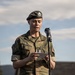 A female military officer in Norwegian Army camouflage uniform is reading from a document at a microphone stand, likely delivering a speech at an outdoor ceremony. She wears a dark beret with a unit badge and a name tape reading 'EIDEM' on her uniform. The setting appears to be a military installation with brick buildings visible in the background.