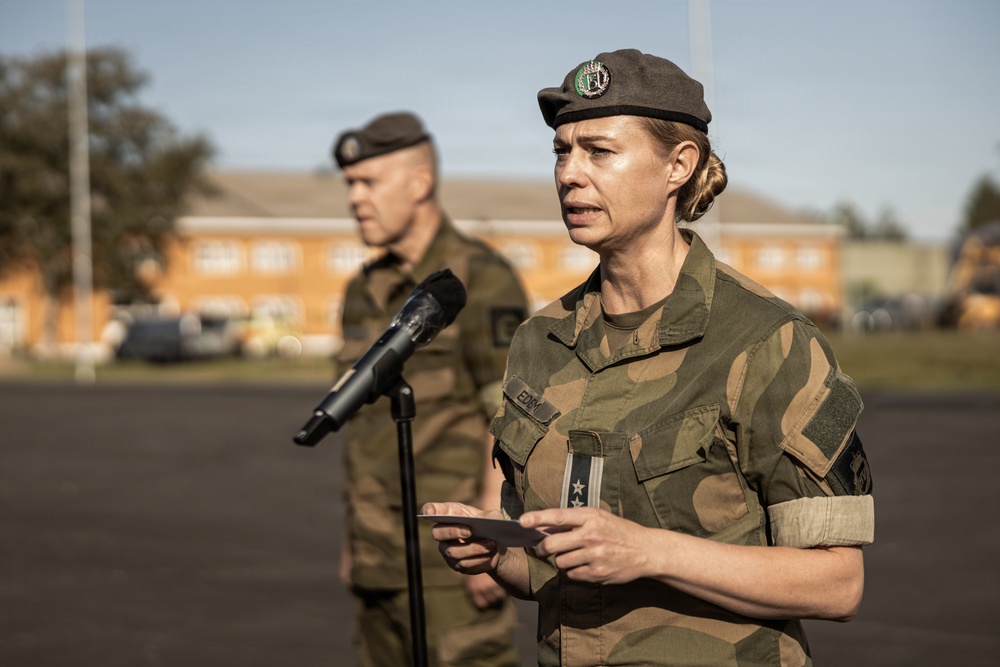 A female military officer in camouflage uniform is speaking at a microphone stand while holding a small card or note. She wears a dark beret with a badge and has two stars visible on her rank insignia. A male soldier in similar uniform stands in the background, slightly out of focus.