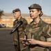 A female military officer in camouflage uniform is speaking at a microphone stand while holding a small card or note. She wears a dark beret with a badge and has two stars visible on her rank insignia. A male soldier in similar uniform stands in the background, slightly out of focus.