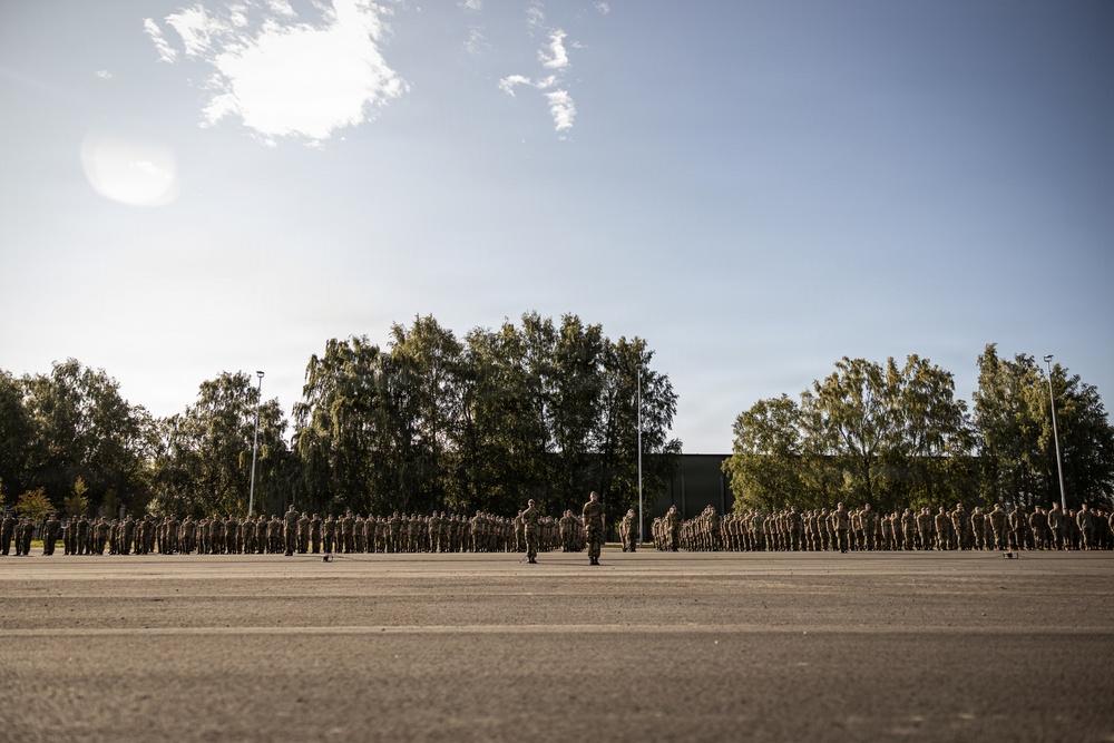 A large military formation is assembled on an open parade ground, with personnel arranged in multiple organized rows facing a central position. Several individuals who appear to be officers or instructors stand in front of the assembled troops. The setting appears to be a military base or training facility surrounded by trees and low buildings.