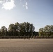 A large military formation is assembled on an open parade ground, with personnel arranged in multiple organized rows facing a central position. Several individuals who appear to be officers or instructors stand in front of the assembled troops. The setting appears to be a military base or training facility surrounded by trees and low buildings.