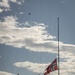 Three flagpoles are visible, with the Norwegian flag flying at half-mast on the tallest pole in the foreground, indicating a period of mourning or commemoration. The middle pole displays the American flag and the left pole displays the British Union Jack, both also appearing to fly at reduced height. The scene is set against a partly cloudy sky with trees visible in the background and a bird in flight near the top of the frame.