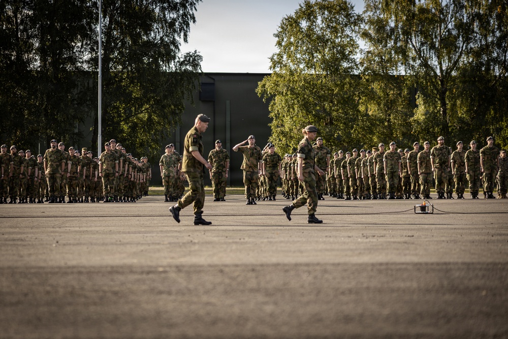 A military parade or formal inspection is taking place on an open parade ground. Two senior personnel are walking in the foreground while a soldier renders a salute, with large formations of uniformed troops standing at attention in the background. The setting appears to be a military base or garrison, likely in a Scandinavian country based on the uniform style and environment.