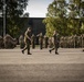 A military parade or formal inspection is taking place on an open parade ground. Two senior personnel are walking in the foreground while a soldier renders a salute, with large formations of uniformed troops standing at attention in the background. The setting appears to be a military base or garrison, likely in a Scandinavian country based on the uniform style and environment.
