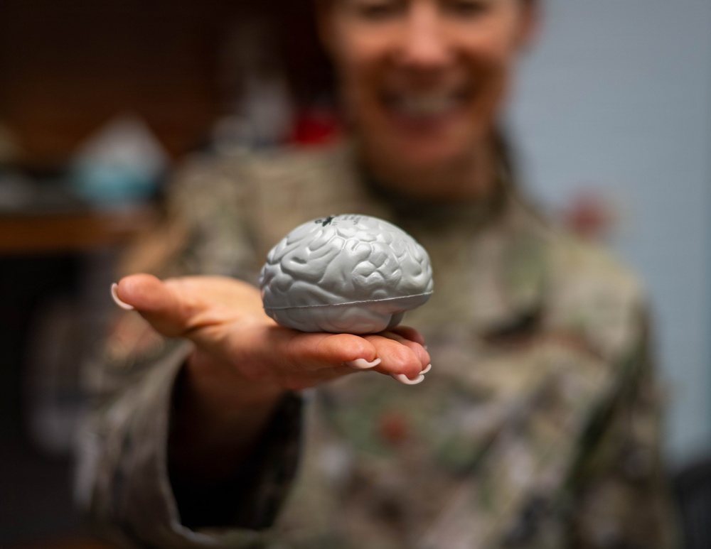A person wearing military camouflage uniform is holding out a small gray brain-shaped object toward the camera on an open palm. The brain model appears to be a novelty item or stress ball with detailed anatomical surface texture. The subject is smiling, and the focus is sharp on the object while the person remains blurred in the background.