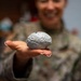 A person wearing military camouflage uniform is holding out a small gray brain-shaped object toward the camera on an open palm. The brain model appears to be a novelty item or stress ball with detailed anatomical surface texture. The subject is smiling, and the focus is sharp on the object while the person remains blurred in the background.