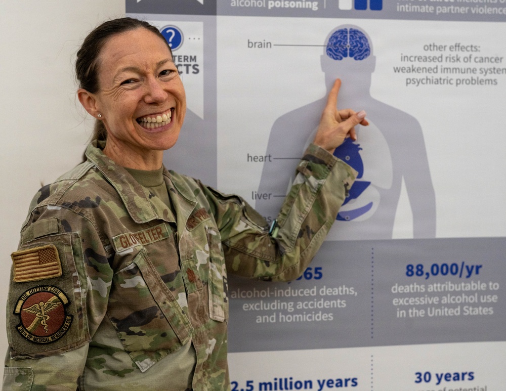 A female military service member in U.S. Army OCP (Operational Camouflage Pattern) uniform is smiling broadly while pointing at an informational poster about the effects of alcohol on the human body. Her name tape reads 'GLOVELTER' and she wears a unit patch identified as '376th OP Medical Readiness SQ' with a caduceus symbol. The poster behind her displays statistics and anatomical diagrams related to alcohol-induced health risks and deaths.